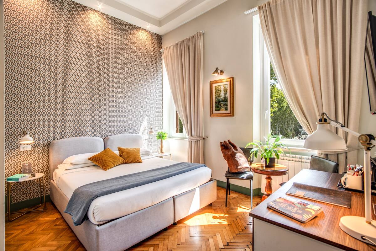 A modern bedroom with a large bed, a desk, a chair, a potted plant, and a window with curtains.