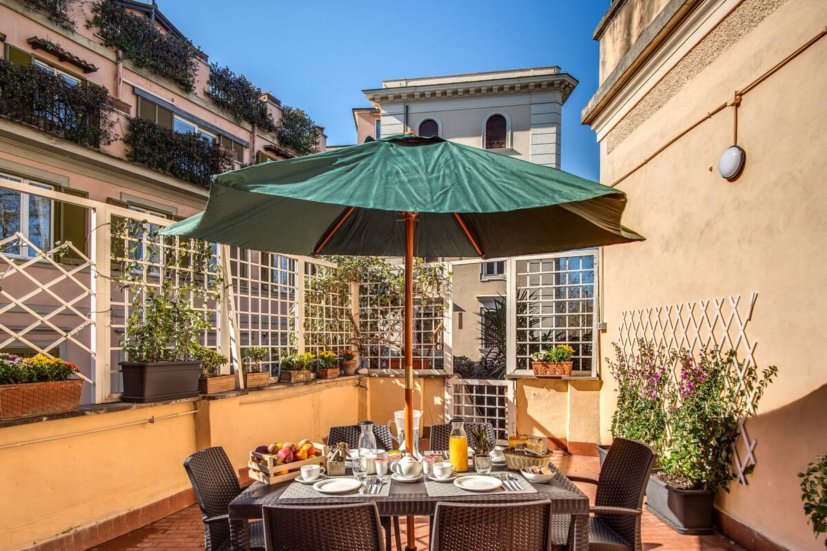 An outdoor dining area with a table set for four, surrounded by wicker chairs, under a green umbrella.