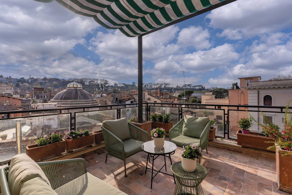 A rooftop terrace with green wicker furniture, potted plants, and a clear view of the cityscape.