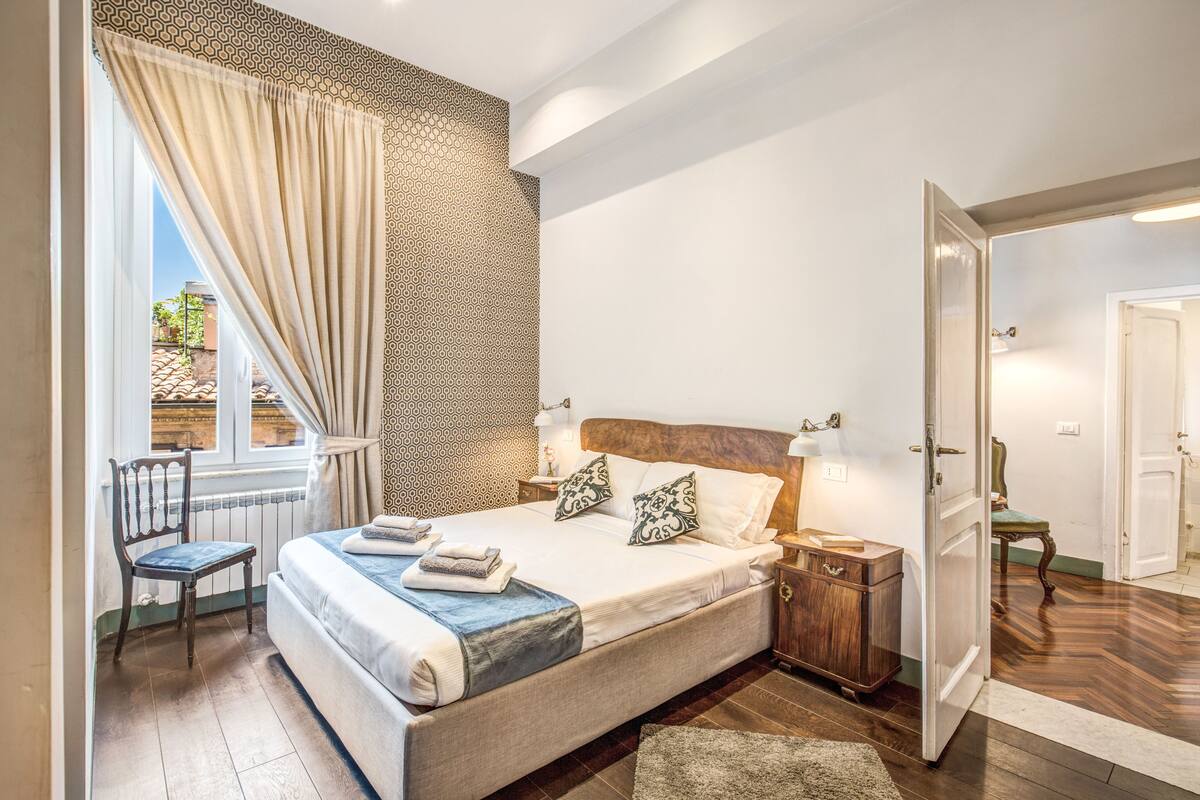A bedroom with a large bed, a chair, a nightstand, and a window with curtains.