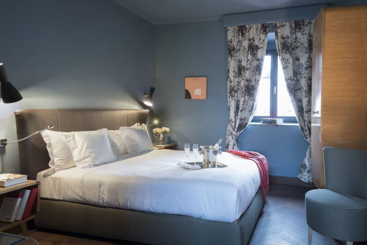 A neatly made bed with white linens, a bedside table with a lamp, a small table with a vase of flowers, and a window with floral curtains.