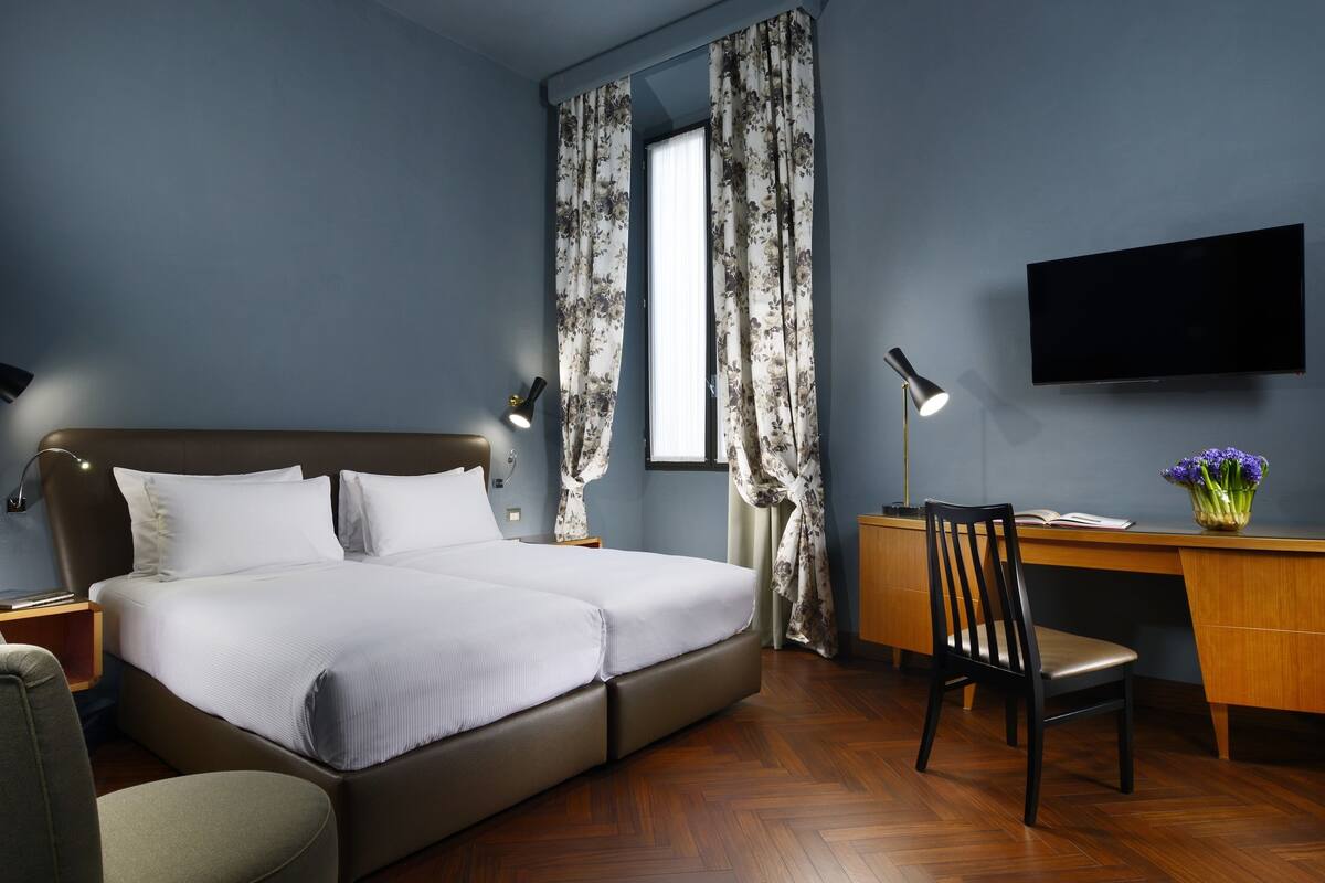 A modern hotel room with a large bed, a desk, a chair, a TV, and a window with curtains.