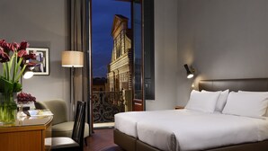 Une chambre d’hôtel avec un lit, un bureau doté d’une lampe, une chaise et une vue sur un immeuble, visible à travers une porte vitrée.