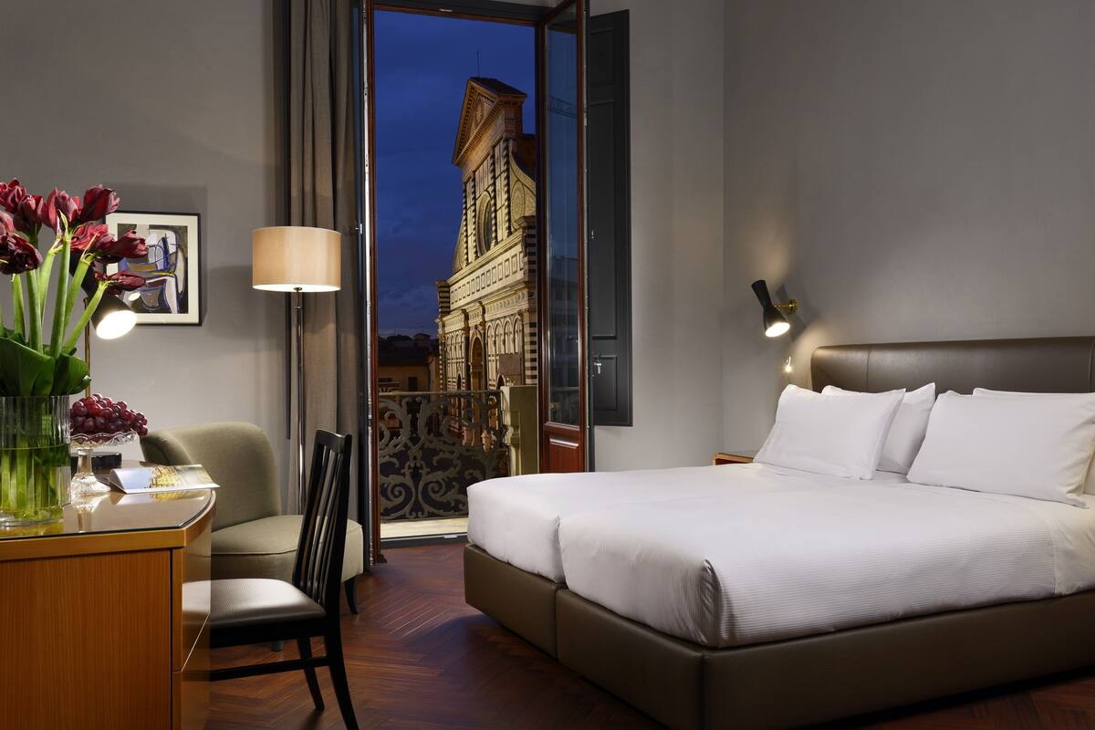 A hotel room with a bed, a desk with a lamp, a chair, and a view of a building through a glass door.