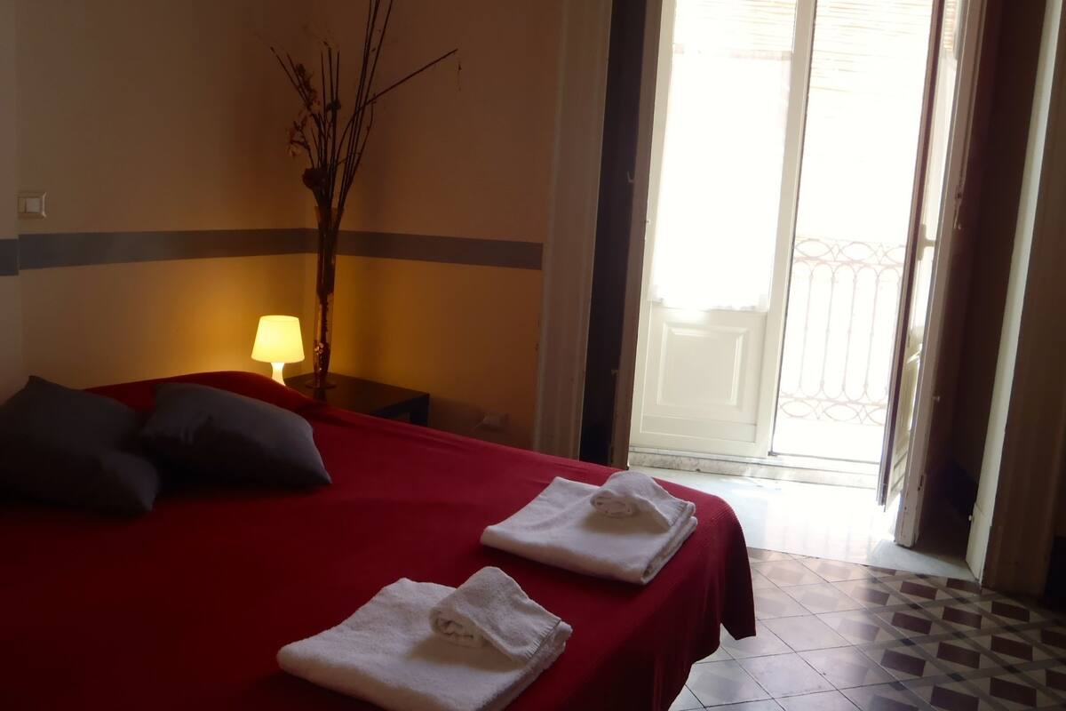 Habitación de hotel con cama, mesita de noche, lámpara, un florero con ramas y vistas al exterior a través de una puerta.