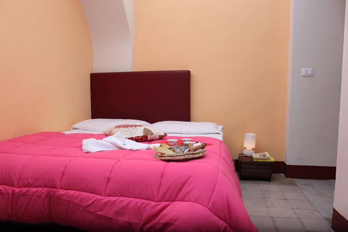 Una cama bien tendida con una colcha rosa brillante, una cesta de pan y una pequeña mesa con una lámpara y un libro.