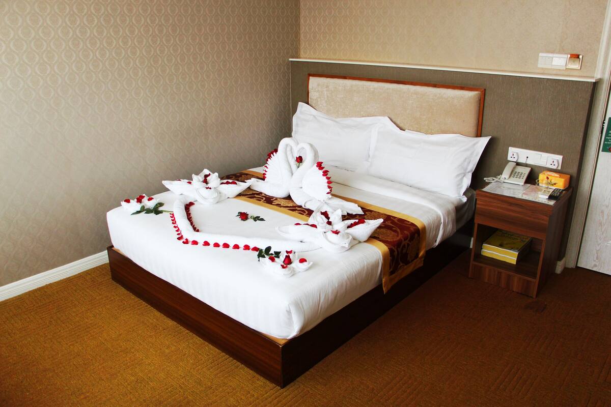 A neatly made bed with white linens and red floral decorations, a wooden headboard, a bedside table with a telephone and a book, and a carpeted floor.