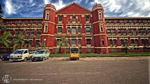 A large, red-brick building with multiple windows and balconies, surrounded by parked cars and palm trees. - Sunny Holiday Hotel (Yangon)