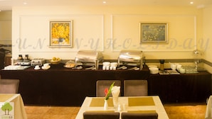 A buffet setup with various food items and utensils, a vase with flowers, and framed artwork on the wall. - Sunny Holiday Hotel (Yangon)