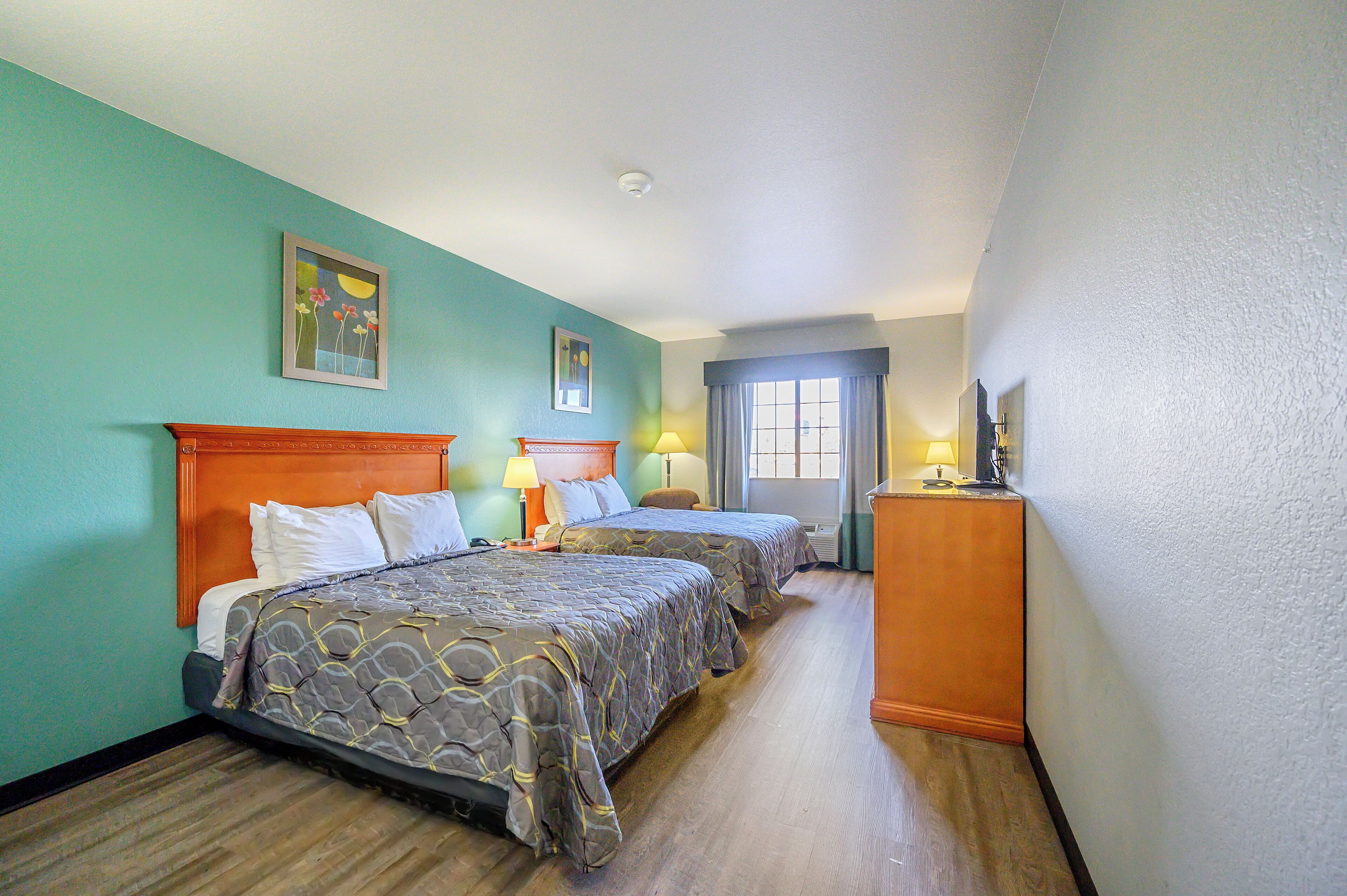 A hotel room with two beds, a wooden headboard, a desk with a lamp, and a window with curtains.