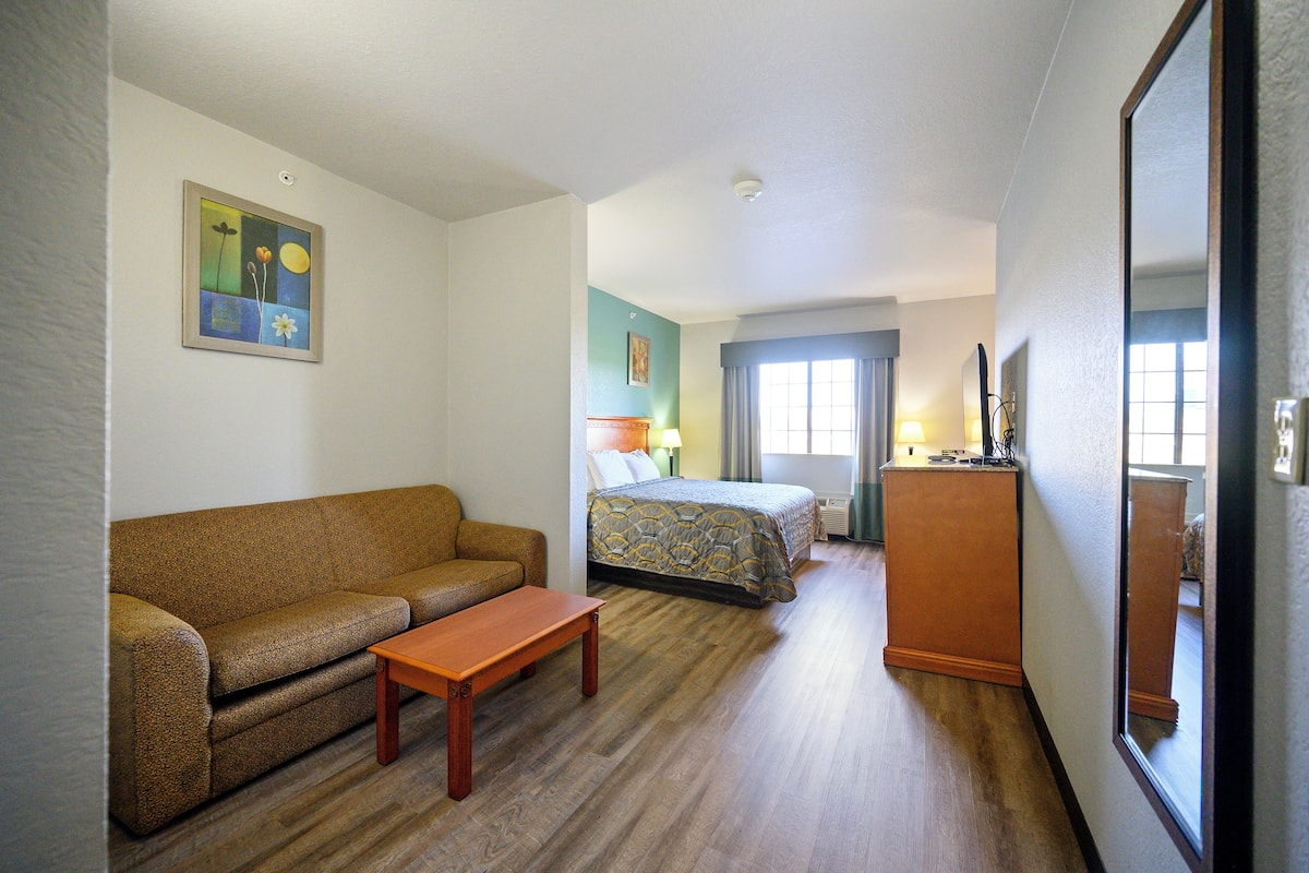 A hotel room with a sofa, a small table, a bed with a patterned bedspread, a nightstand with a lamp, and a large mirror.