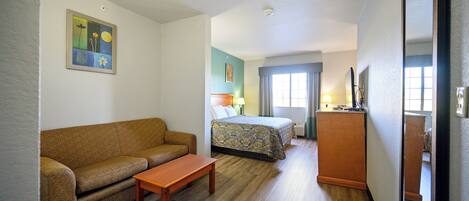 A hotel room with a sofa, a small table, a bed with a patterned bedspread, a nightstand with a lamp, and a large mirror.
