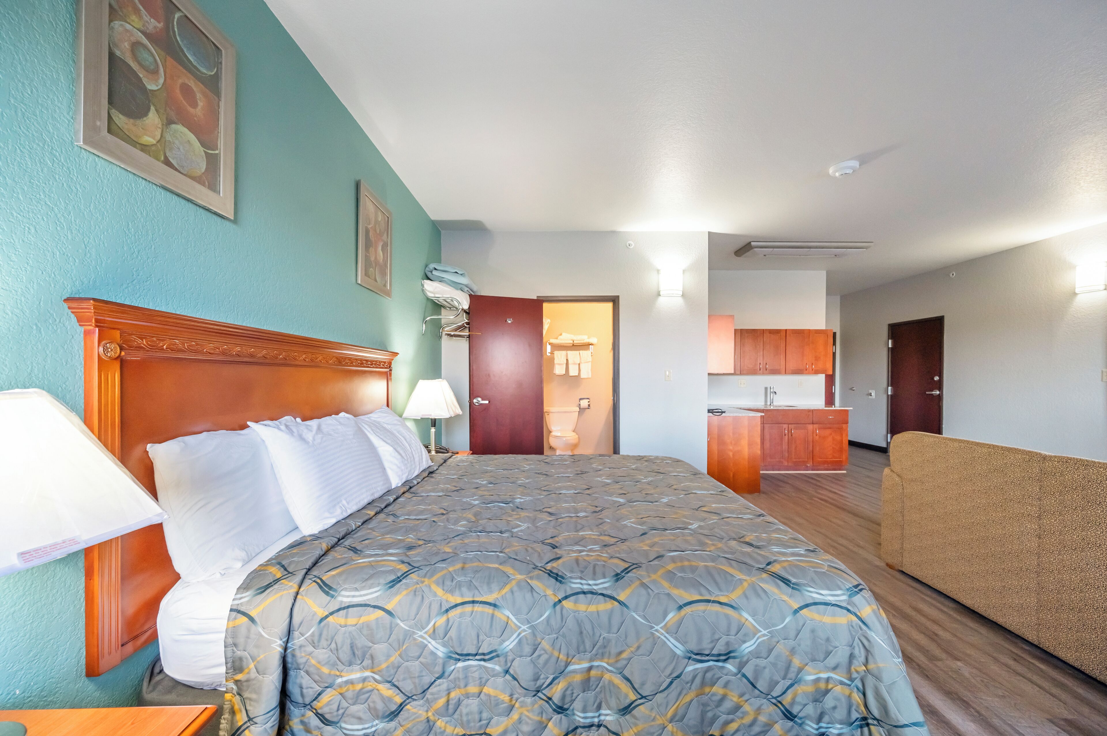 Habitación de hotel con una cama grande, cabecera de madera, sofá, kitchenette y baño visible a través de una puerta abierta.