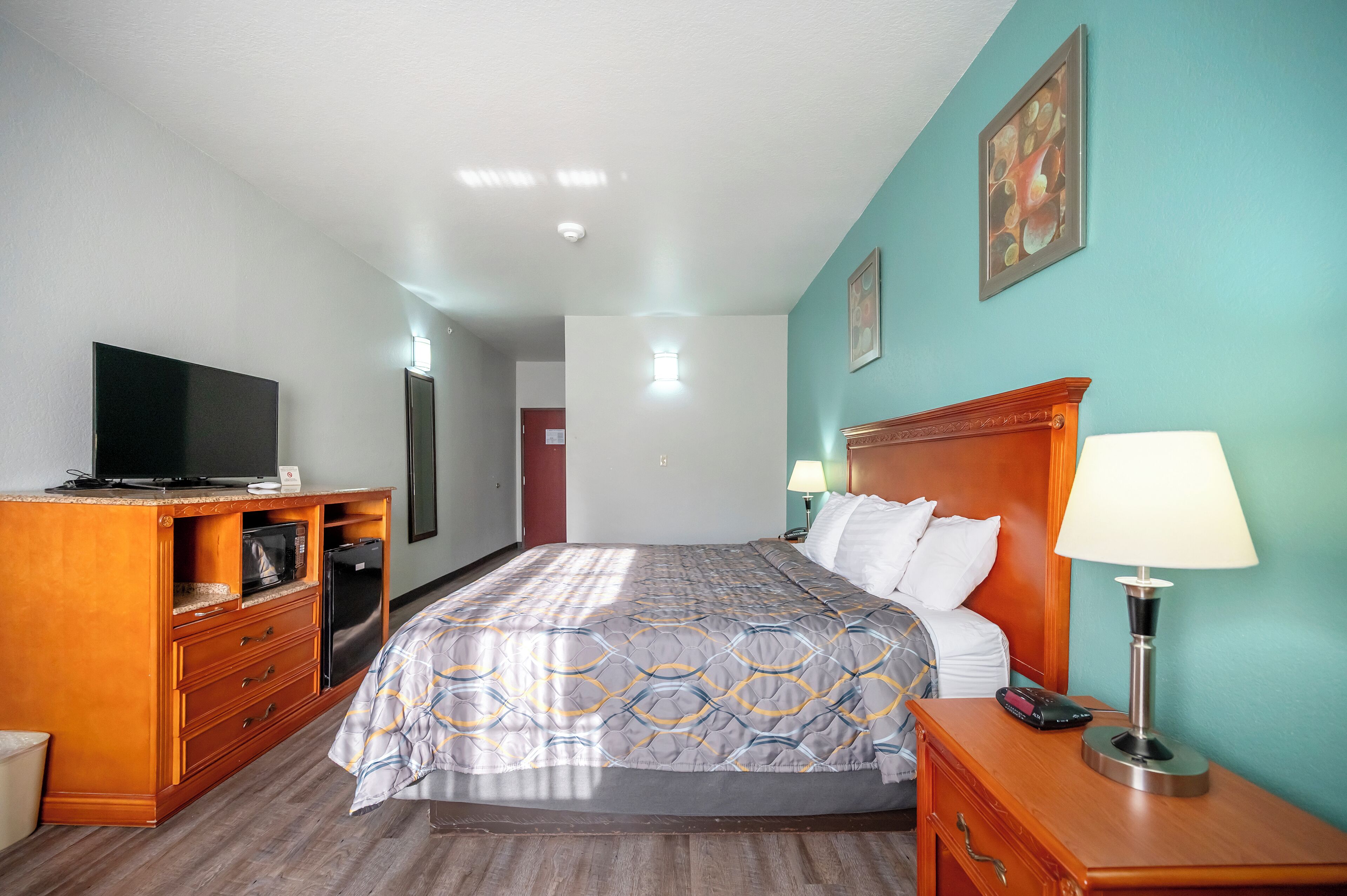 A hotel room with a bed, a TV, a wooden dresser, and a nightstand with a lamp.