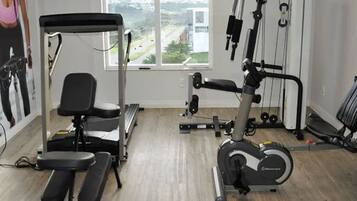 A gym with exercise equipment, a bench, and a window with a view of a road and greenery.