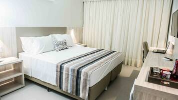 Una habitación de hotel con cama, mesitas de noche, un escritorio con una computadora portátil, una silla y un amplio ventanal con cortinas.