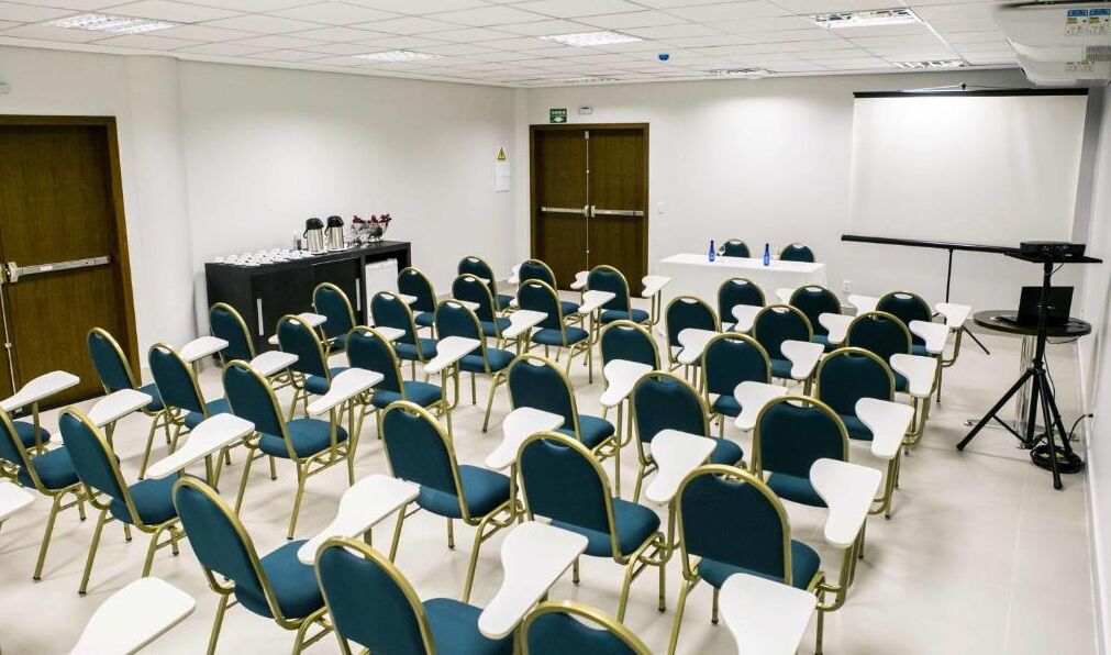 Un salón de conferencias con filas de sillas azules y mesas blancas, un proyector y una pantalla.