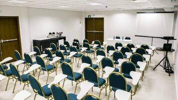 Un salón de conferencias con filas de sillas azules y mesas blancas, un proyector y una pantalla.