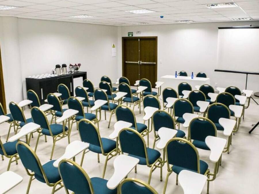Ein Konferenzraum mit Reihen aus blauen Stühlen und weißen Tischen, einem Projektor und einer Leinwand.