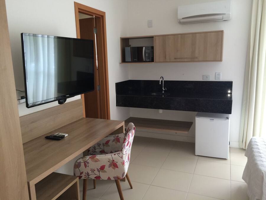 A compact room with a wooden desk, a chair, a flat-screen TV, a small refrigerator, a black countertop with a sink, and a microwave.