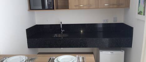 Cozinha compacta com bancada preta, micro-ondas embutido e uma geladeira pequena.