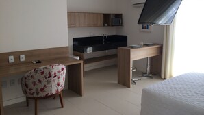 Una habitación de hotel con cama, escritorio con silla, kitchenette y televisor en la pared.