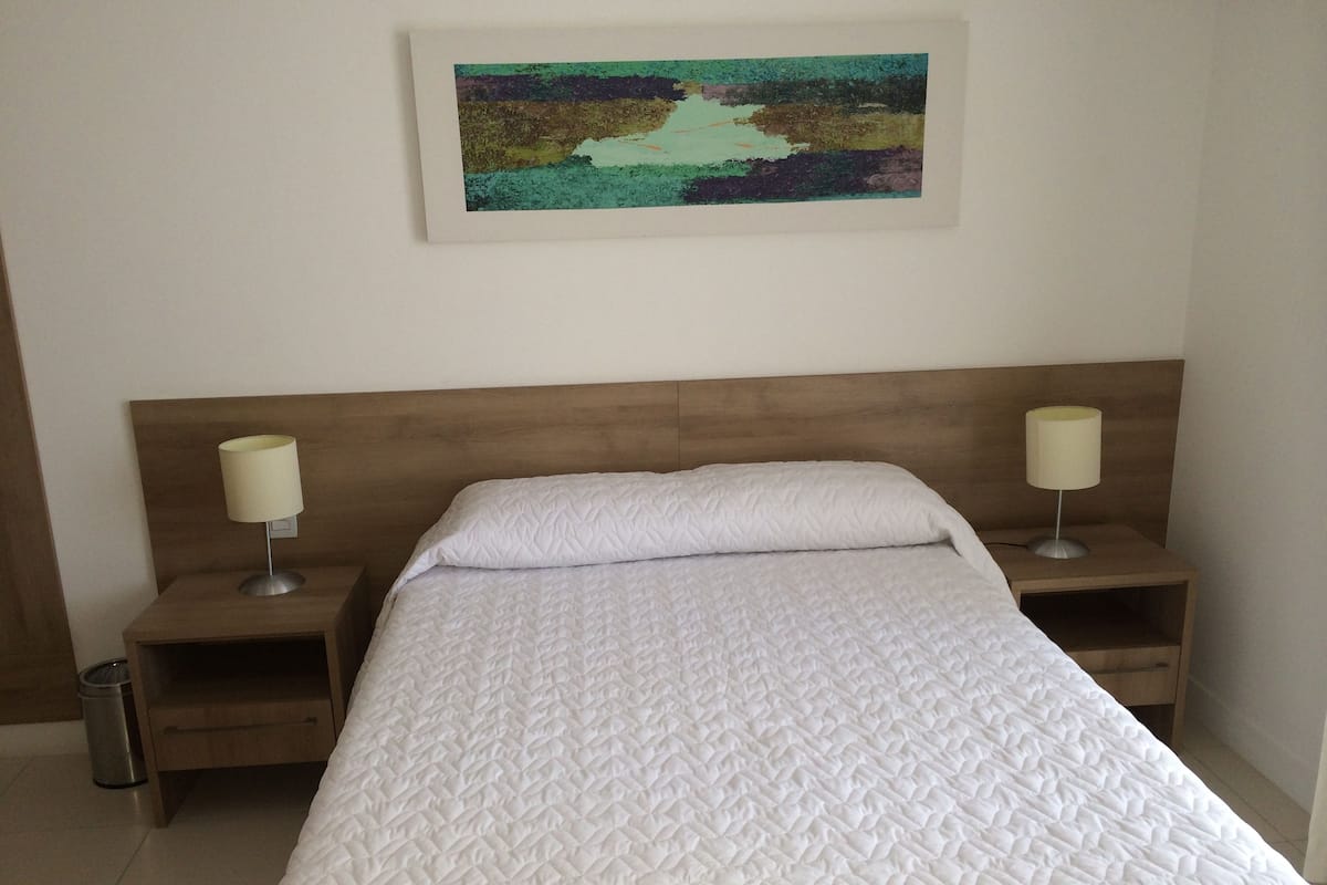 Una cama bien hecha con una colcha blanca acolchada, dos lámparas de noche y un cuadro abstracto enmarcado en la pared.