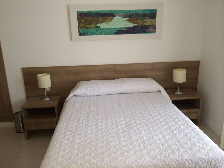 A neatly made bed with a white quilted bedspread, two bedside lamps, and a framed abstract painting on the wall.