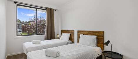 Habitación de hotel con dos camas, cabecero de madera, una mesita con lámpara y una ventana con cortinas.