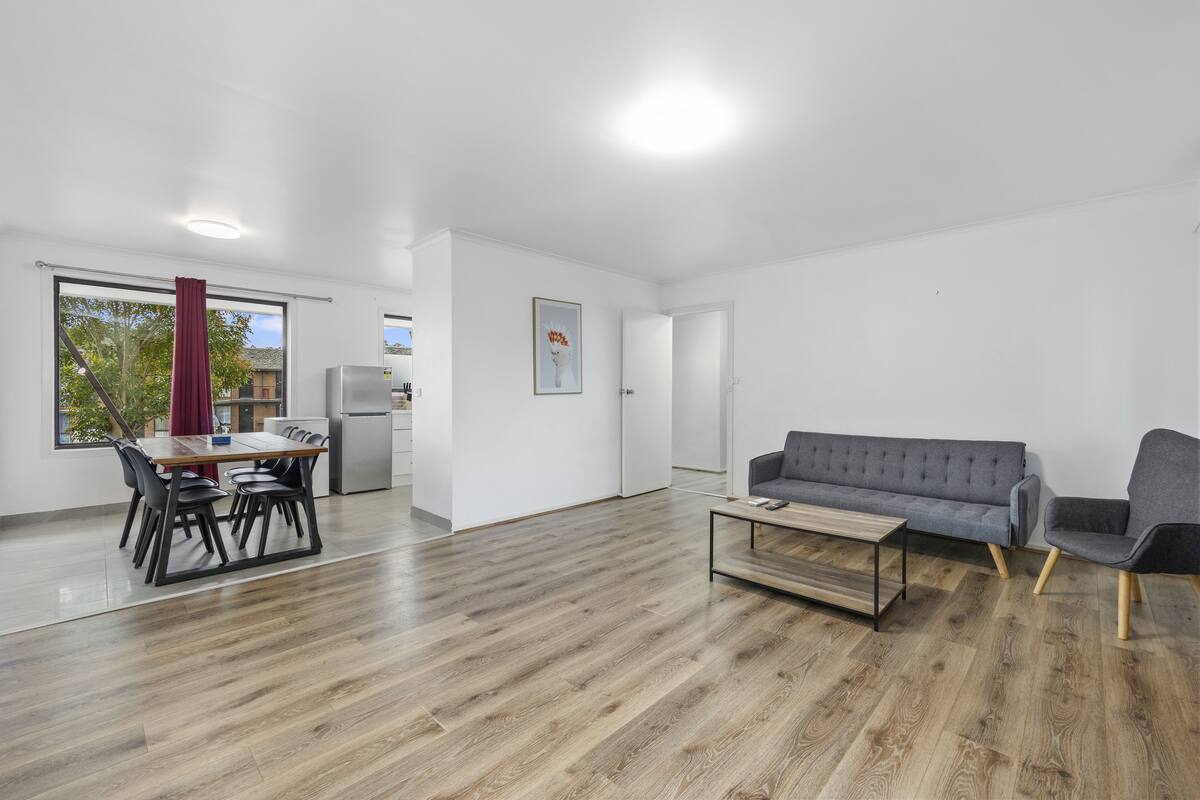 Ein modernes Wohnzimmer mit Holzboden, einem grauen Sofa, einem Esstisch mit Stühlen und einer Küche im Hintergrund.