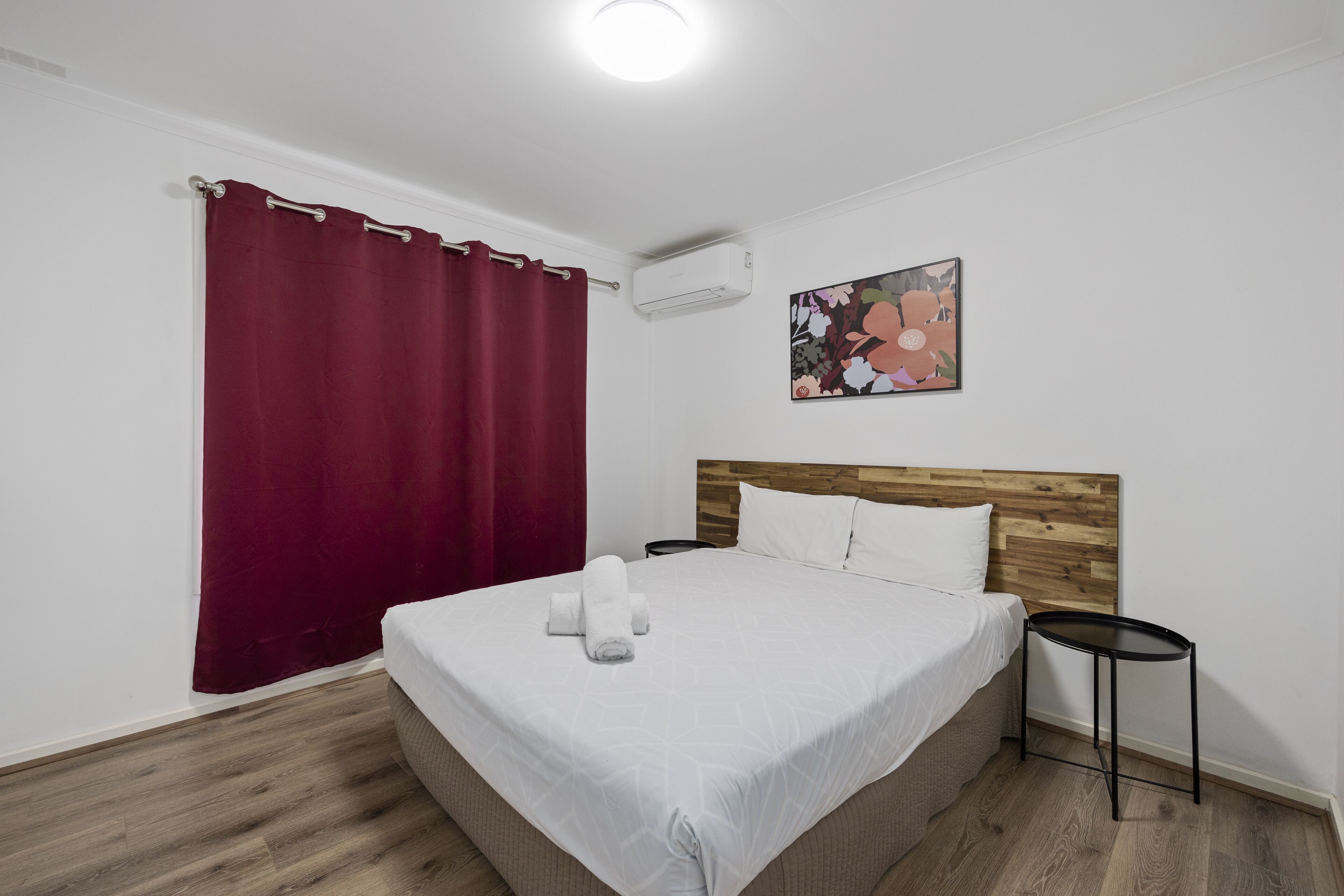 A neatly made bed with a white bedspread and two pillows, a wooden headboard, a small black side table, a red curtain, and a wall-mounted air conditioner.