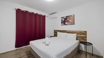 A neatly made bed with a white bedspread and two pillows, a wooden headboard, a small black side table, a red curtain, and a wall-mounted air conditioner.