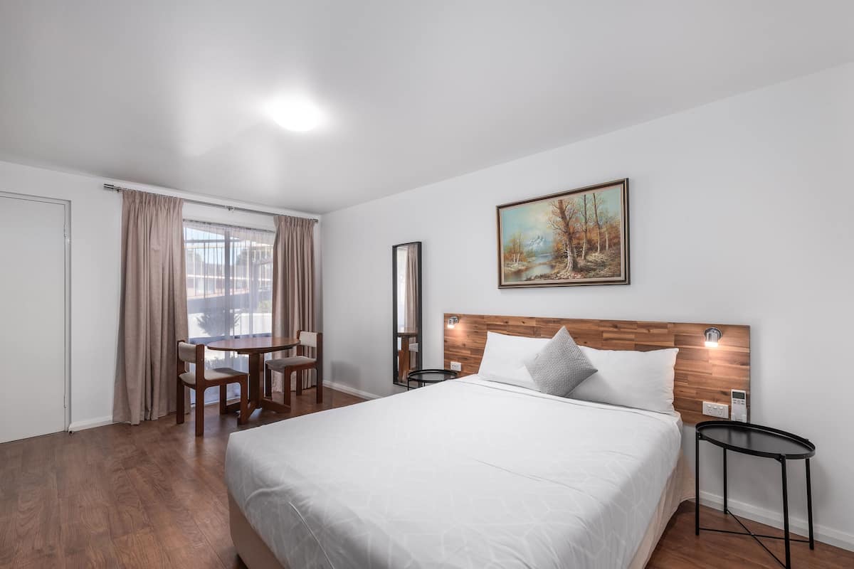 Ein Hotelzimmer mit einem großen Bett, einem hölzernen Kopfteil, einem Gemälde an der Wand, einem Esstisch mit Stühlen und Blick aus dem Fenster.