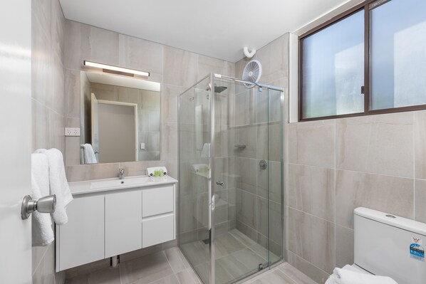 Un baño moderno con ducha de vidrio, tocador blanco con espejo y un inodoro con una etiqueta visible.