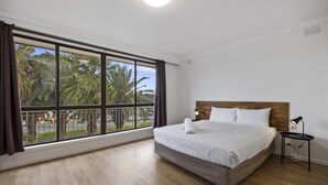 Habitación de hotel con un ventanal amplio, una cama, una mesita de noche y vistas a palmeras y una calle.