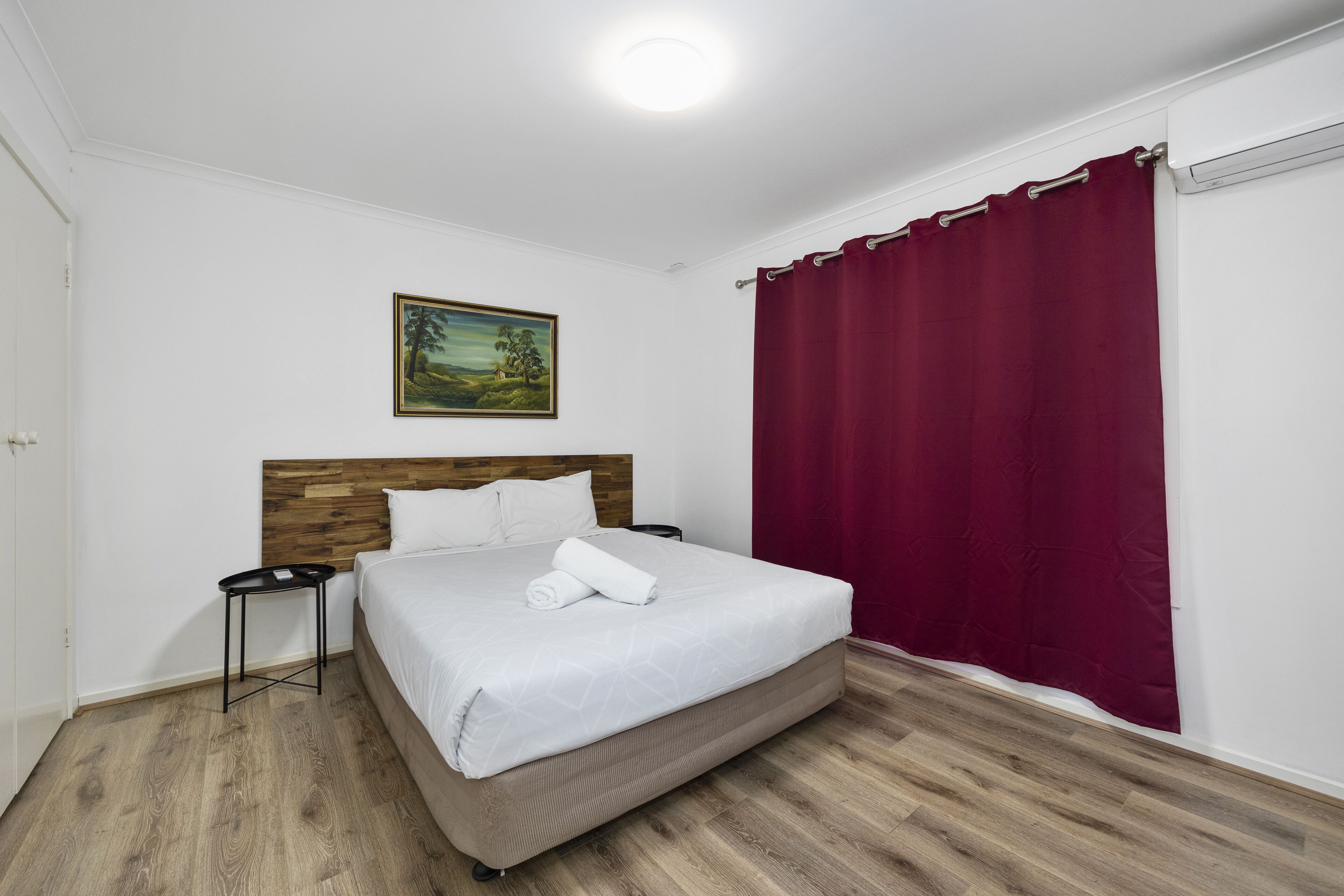 A neatly arranged bedroom with a wooden headboard, a single bed with a white comforter, a small bedside table, and a red curtain.