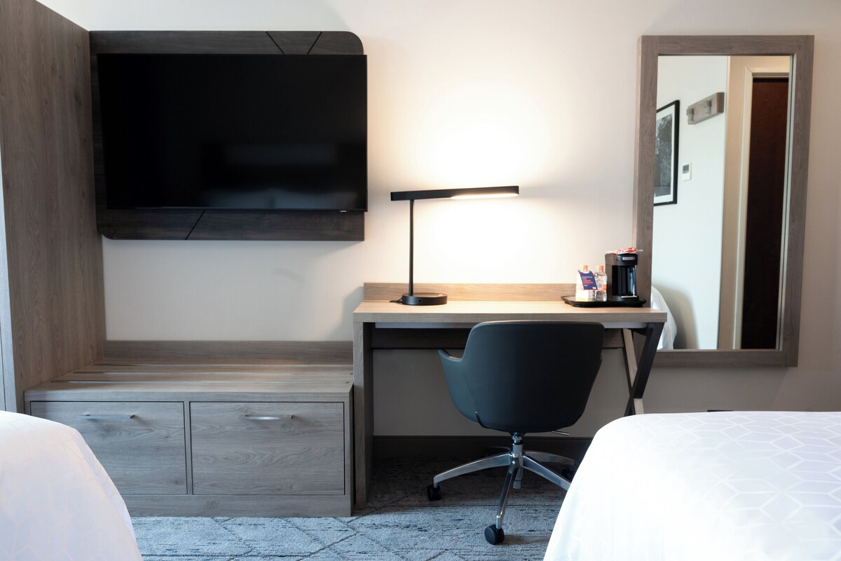 A hotel room with a bed, a desk with a chair, a flat-screen TV mounted on the wall, a desk lamp, and a coffee maker.