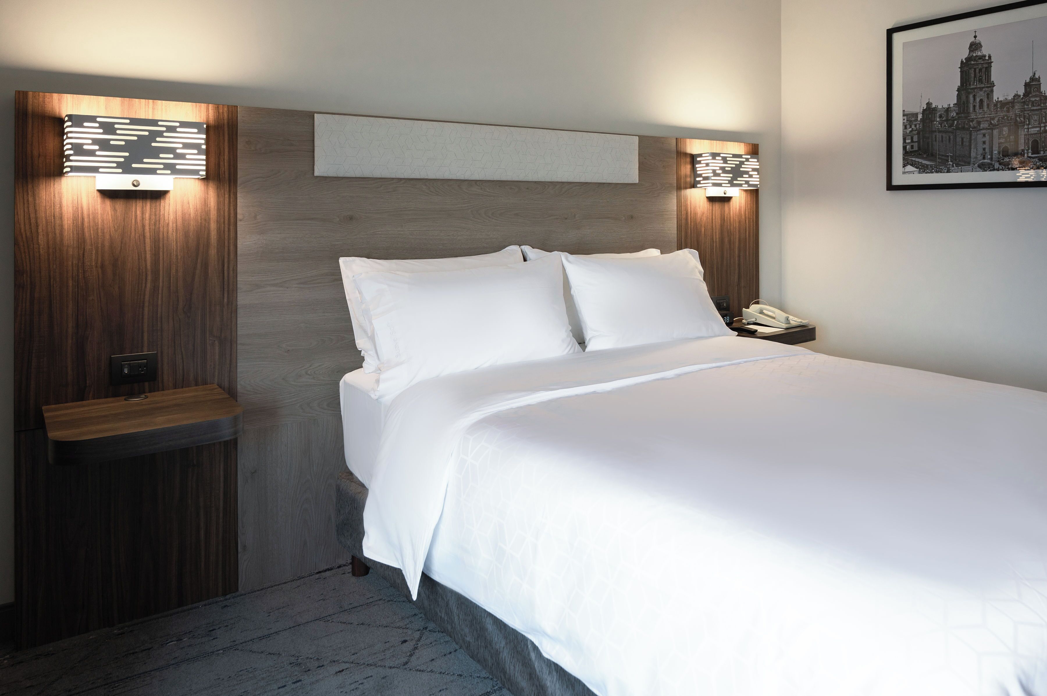 A hotel room with a large bed, wooden headboard, bedside tables, and a framed picture on the wall.