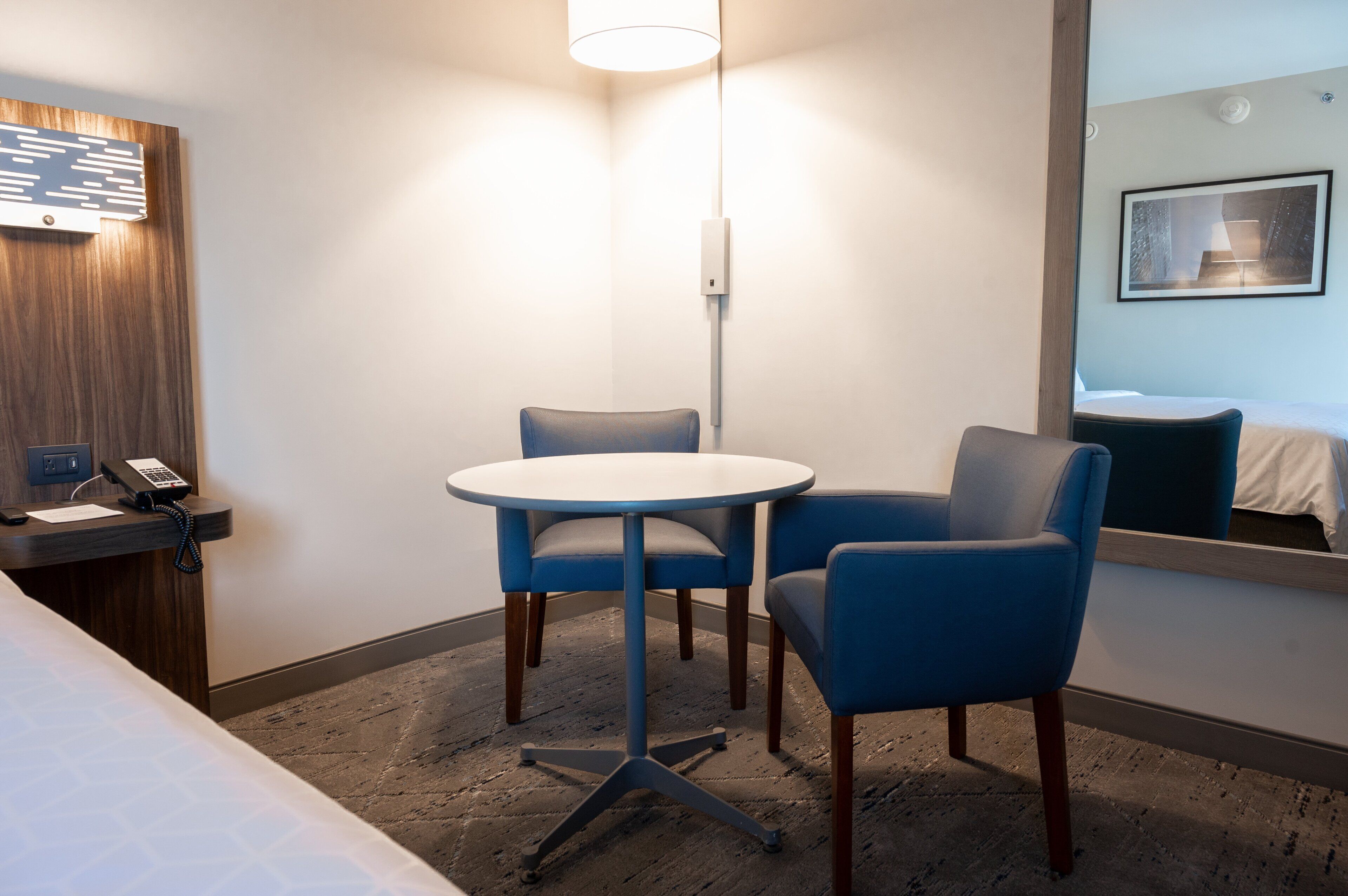 Une chambre d’hôtel comprenant un lit, un bureau avec un téléphone, une table ronde avec deux chaises, un luminaire fixé au mur et un miroir qui reflète le lit et un tableau accroché au mur.