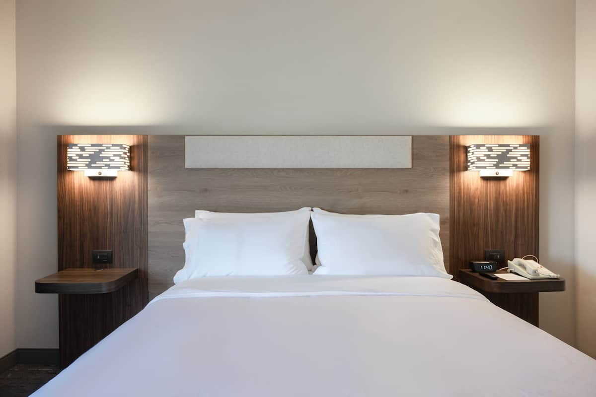 A hotel room with a large bed, two bedside tables, a nightstand, a wall-mounted light fixture, and a clock.