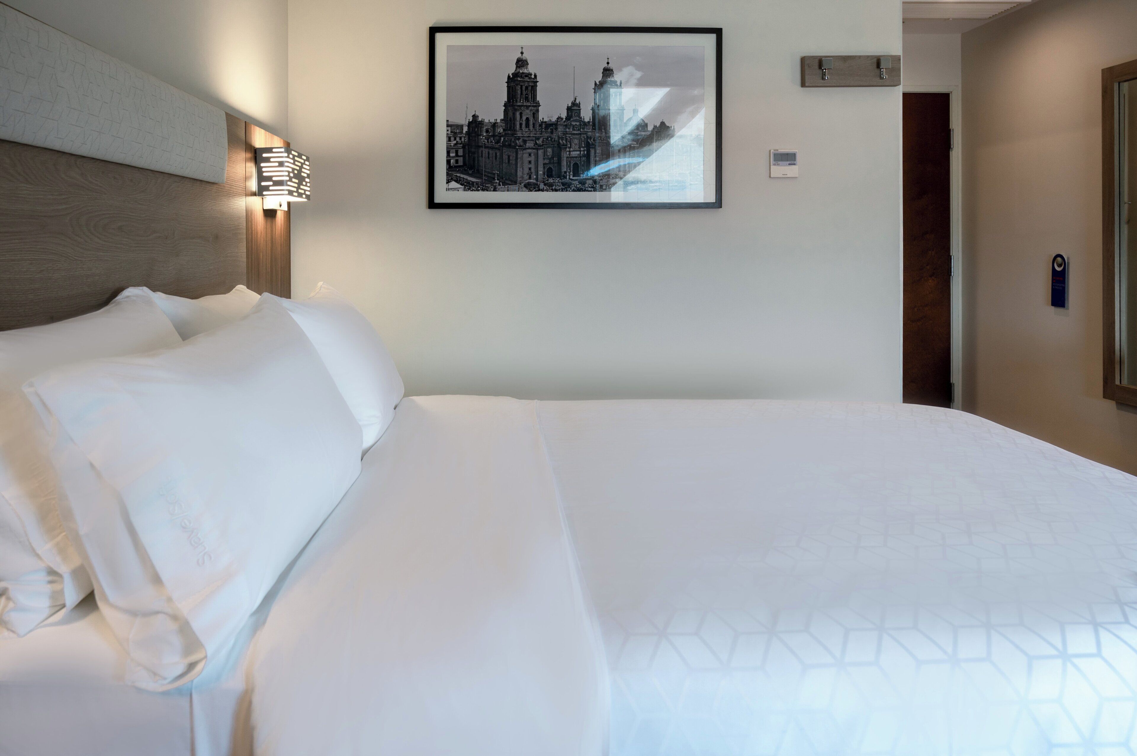 A hotel room with a large bed, a framed picture of a building, a thermostat, and a door with a keycard lock.