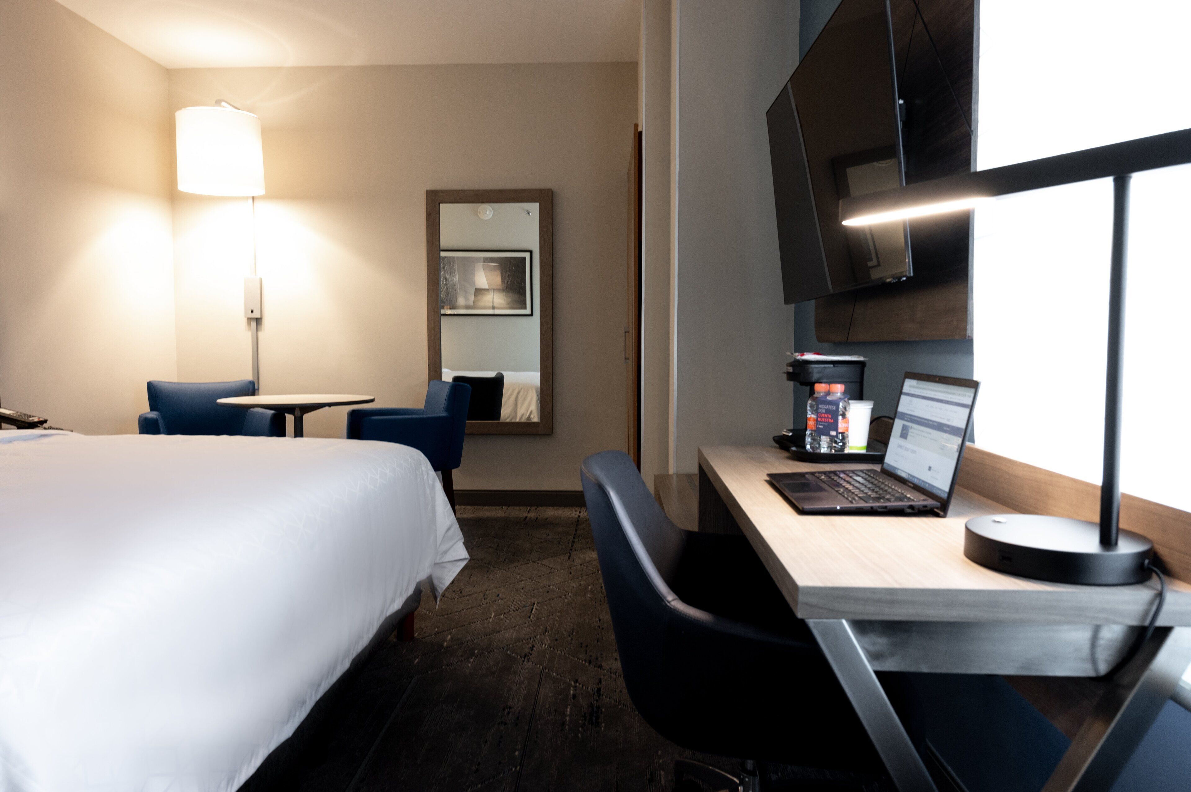 A hotel room with a bed, a desk with a laptop, a chair, a lamp, and a coffee machine.