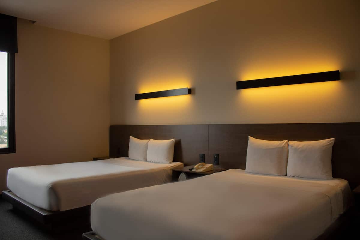 A hotel room with two beds, a nightstand with a telephone, and wall-mounted lighting.