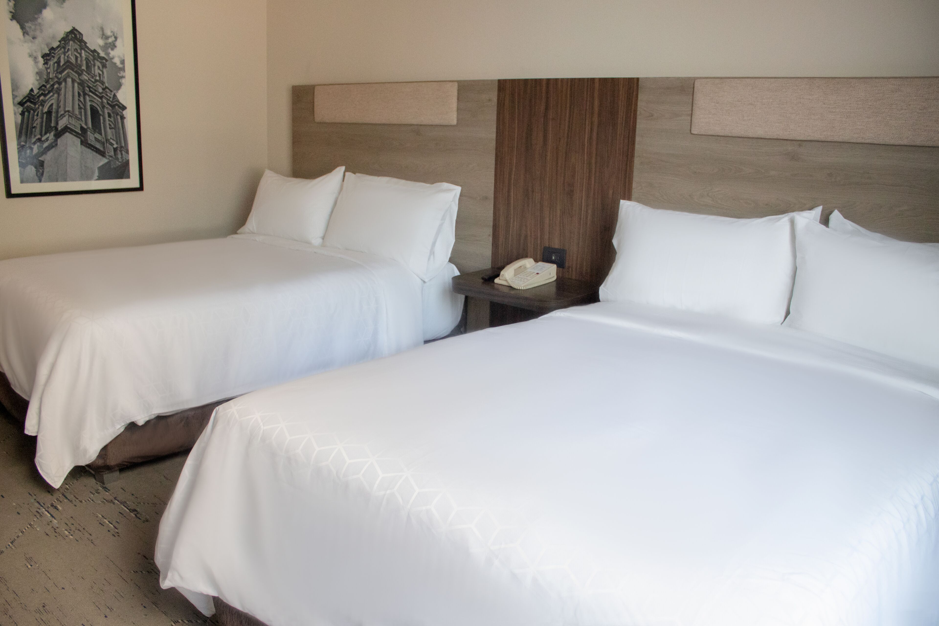 A hotel room with two beds, a nightstand with a phone, and a framed picture on the wall.