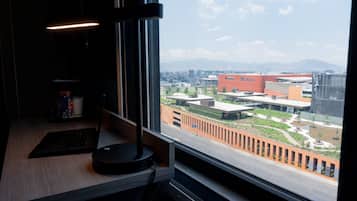 A desk with a laptop and a lamp, a window with a view of buildings and a landscaped area.