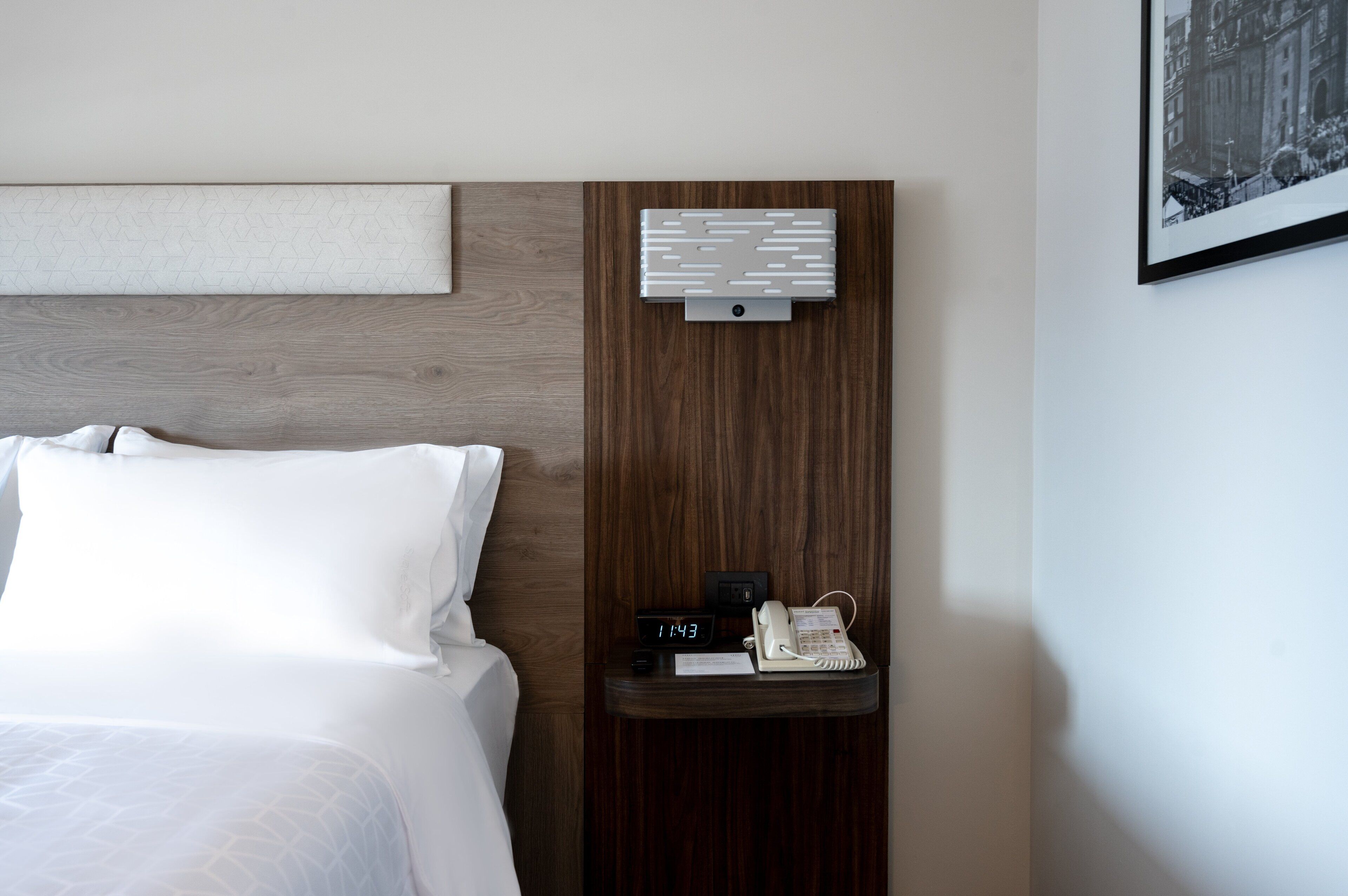 Una habitación de hotel con una cama, una mesita de noche con un reloj y una lámpara fijada a la pared.