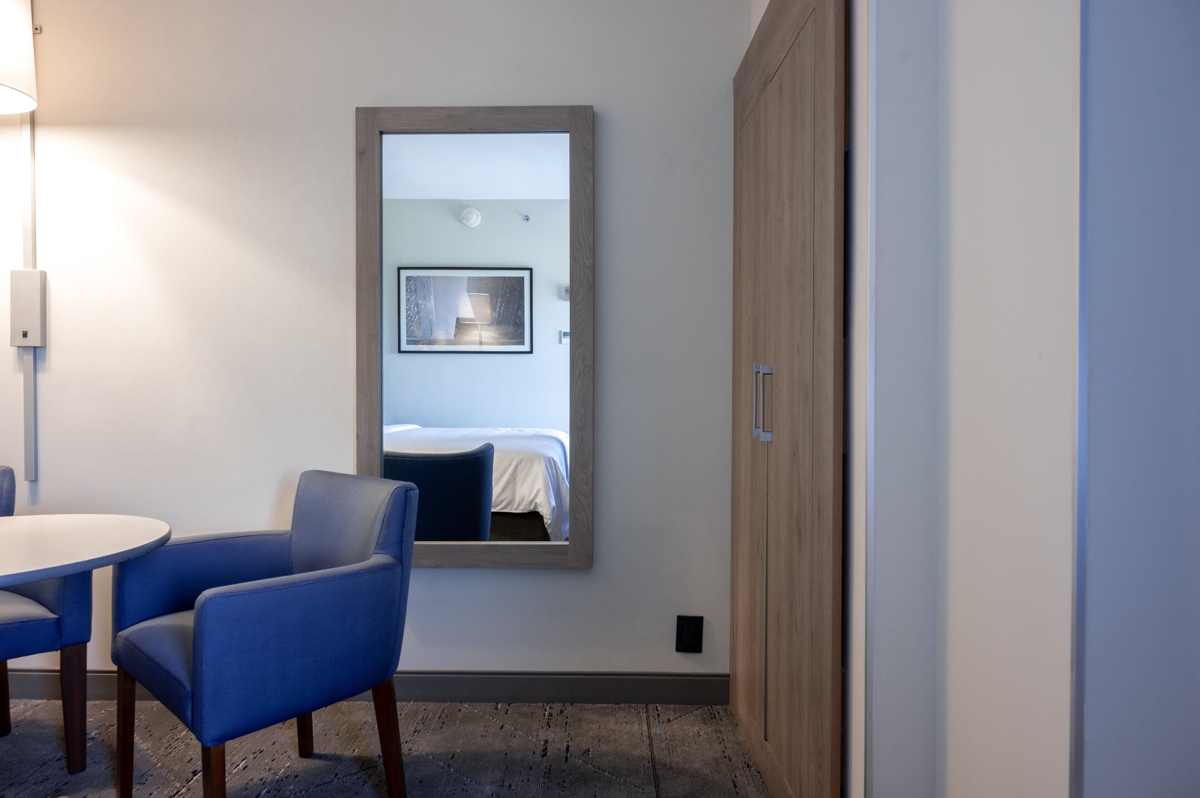 A hotel room with a round table, two blue chairs, a wooden wardrobe, and a large mirror reflecting a bed and a lamp.