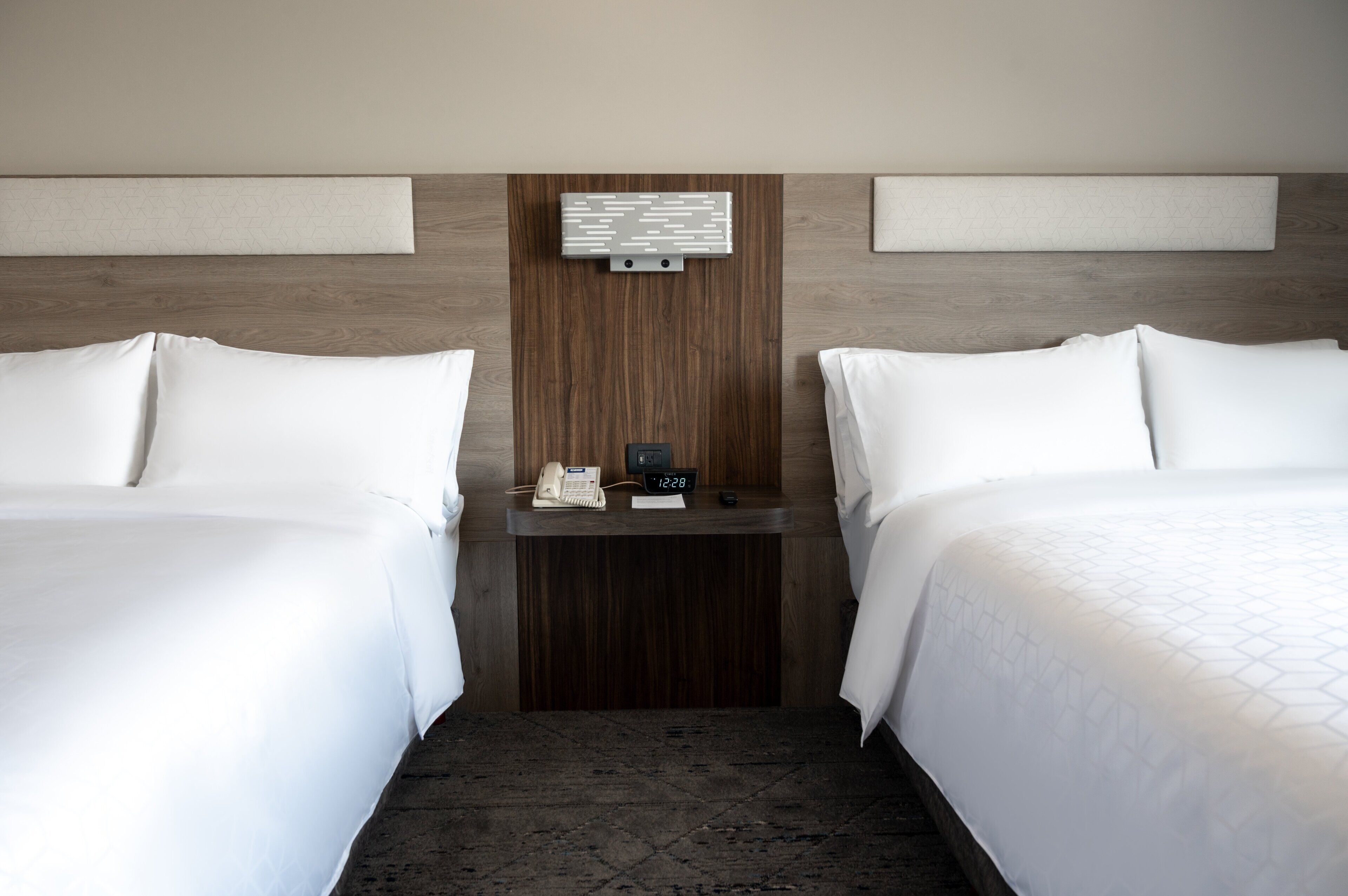 A hotel room with two beds, a wooden headboard, a bedside table with a phone and clock, and a wall-mounted light fixture.