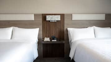 A hotel room with two beds, a wooden headboard, a bedside table with a phone and clock, and a wall-mounted light fixture.