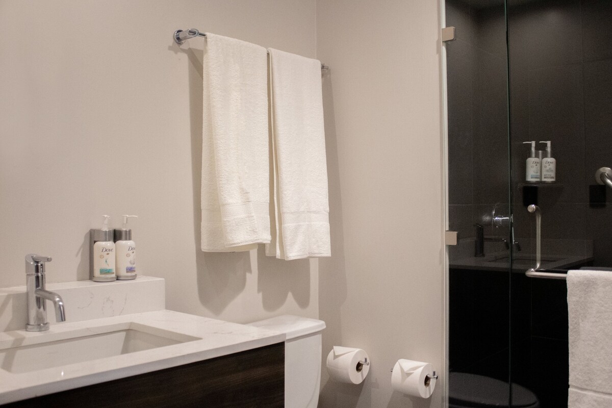 A bathroom with a white sink, two white towels, a glass shower enclosure, and toiletries on the counter.